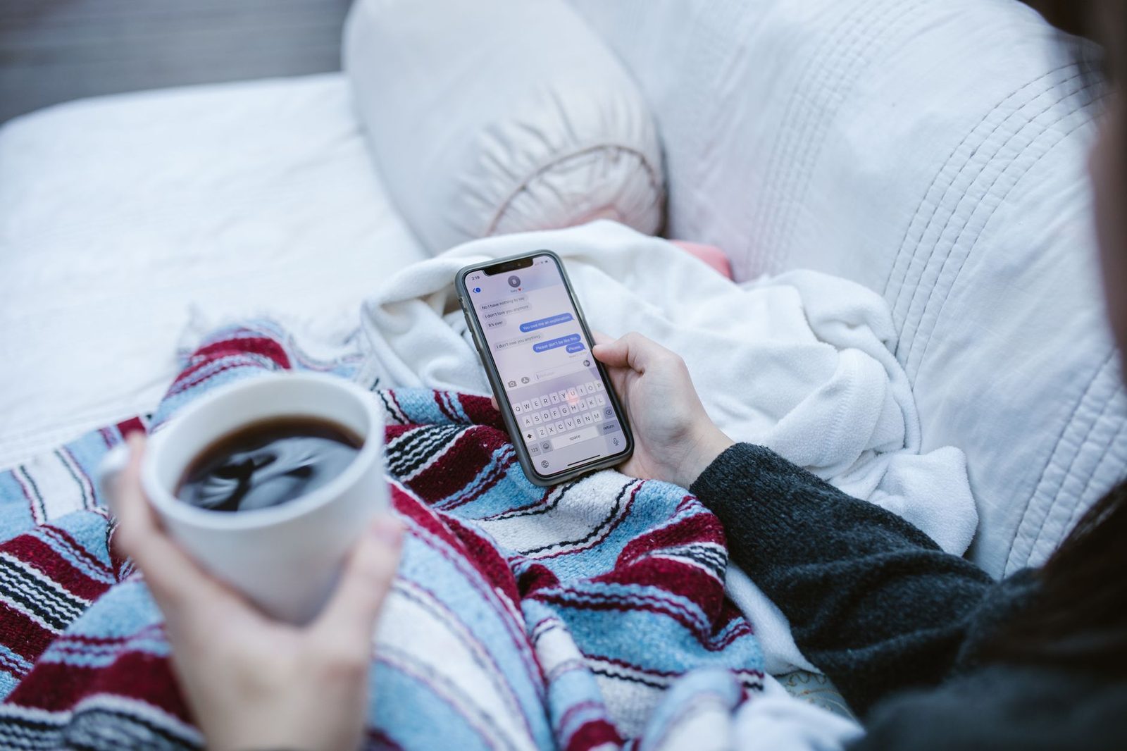 Frau liegt unter einer Decke und liest SMS auf ihrem Smartphone, eine Tasse Kaffee in der Hand