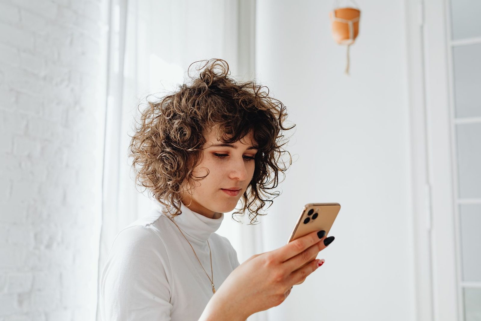 Junge Frau mit lockigem Haar liest vor einem hellen Fenster eine Nachricht auf ihrem Mobiltelefon