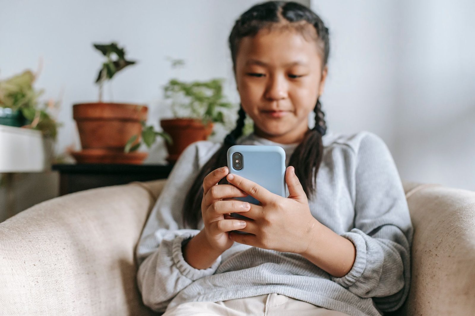 Jeune fille assise dans un canapé, tenant un smartphone bleu entre ses mains, plantes en arrière-plan