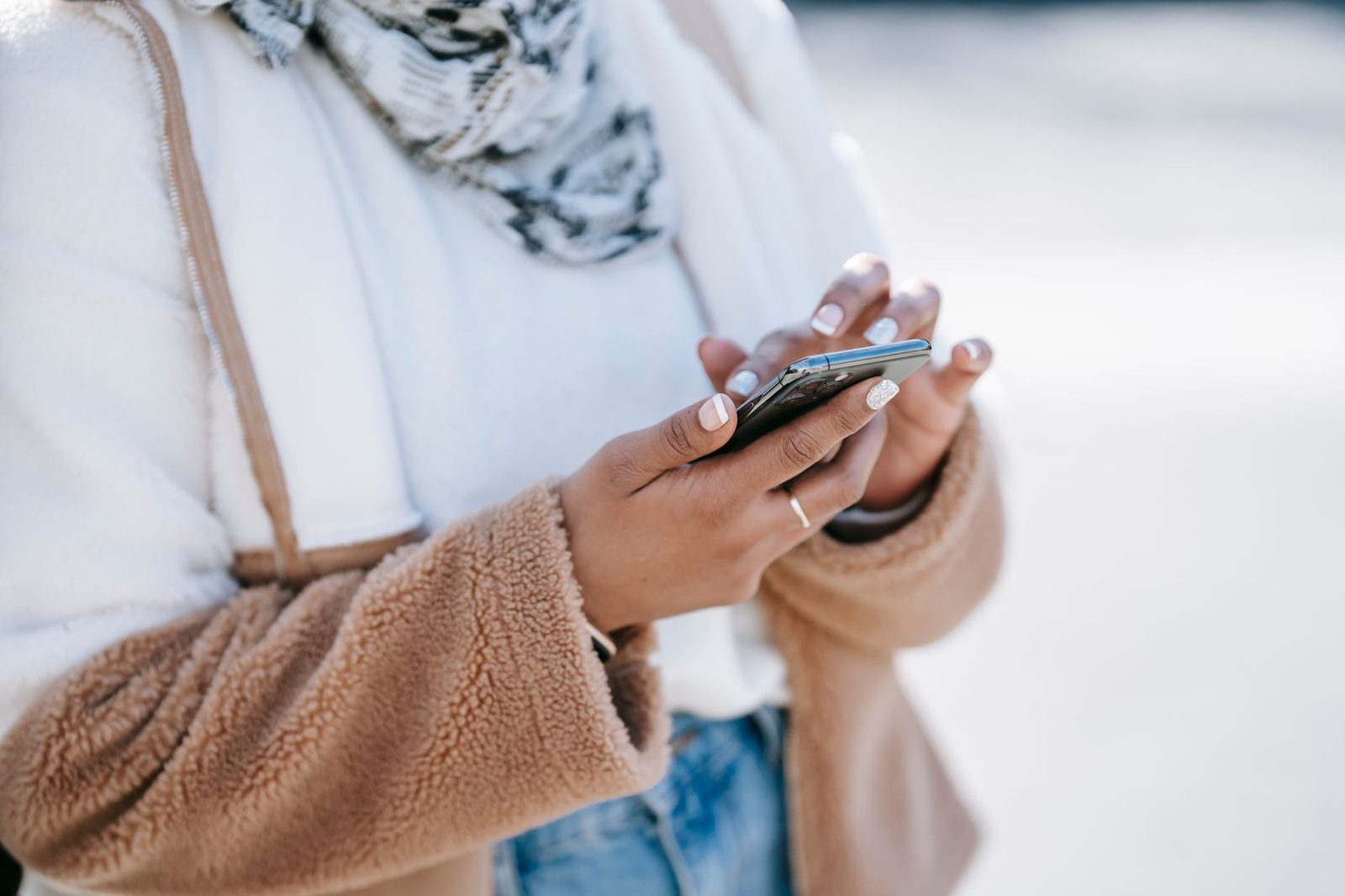 Close-up of the hands of a woman in a beige coat holding and looking at a smartphone outdoors