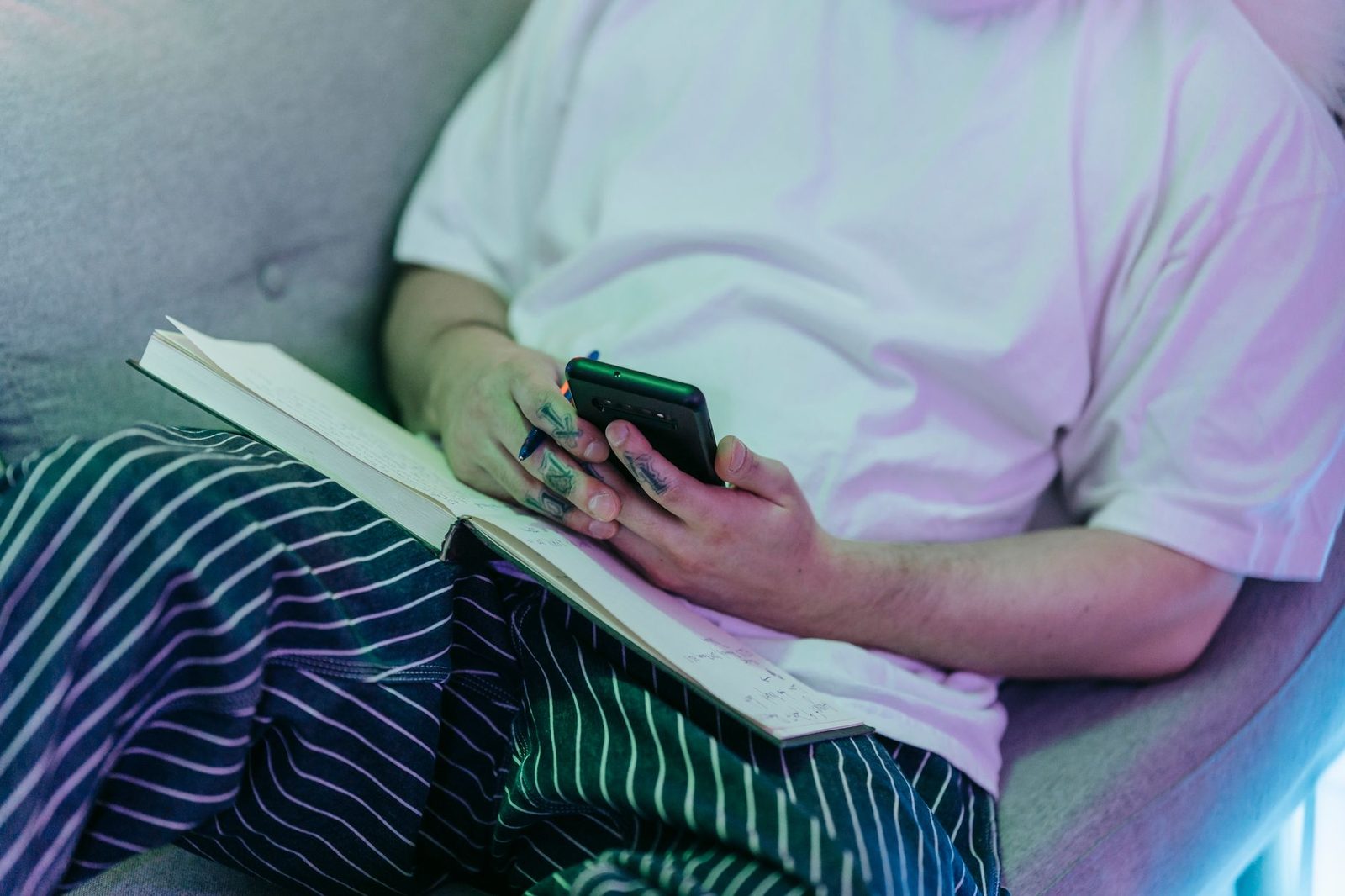 Personne assise sur un canapé, un carnet ouvert sur les genoux, consultant un smartphone tenu à la main