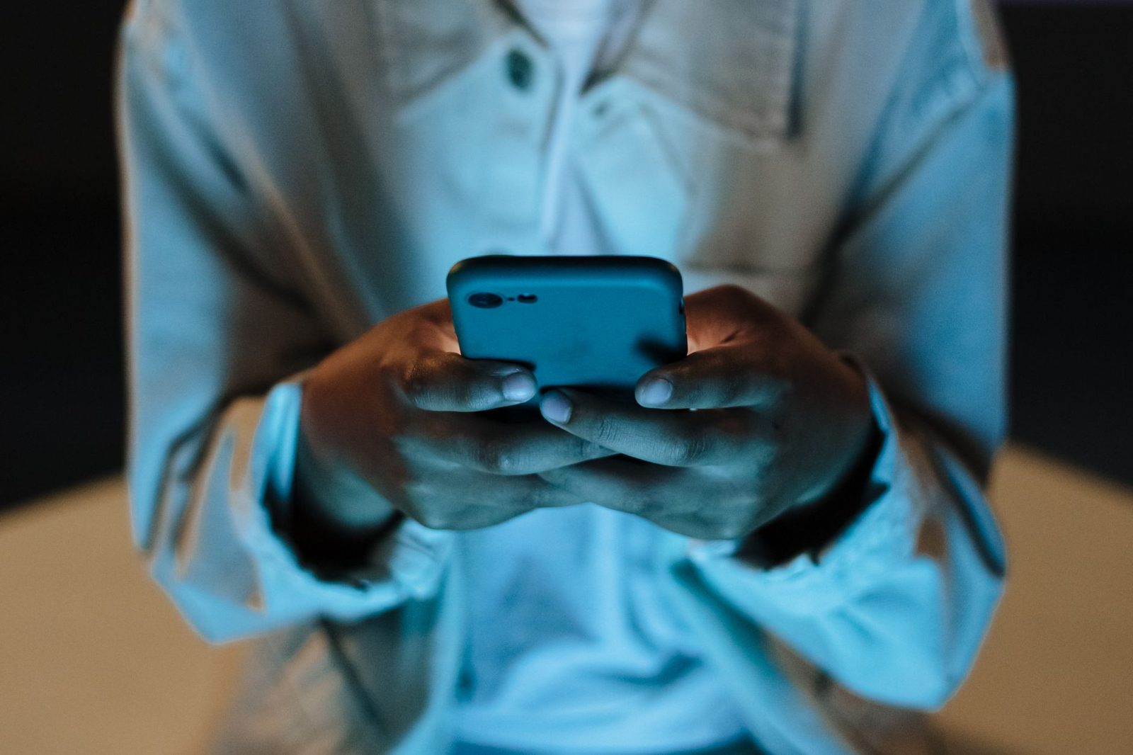 Personne tenant un smartphone à deux mains dans la pénombre, visage hors champ, écran éclairant sa chemise