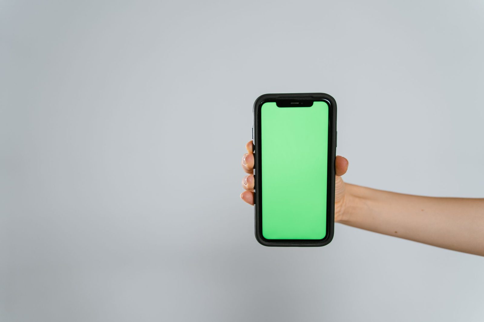 Dłoń trzymająca czarny smartfon z pustym zielonym ekranem na jasnoszarym tle
