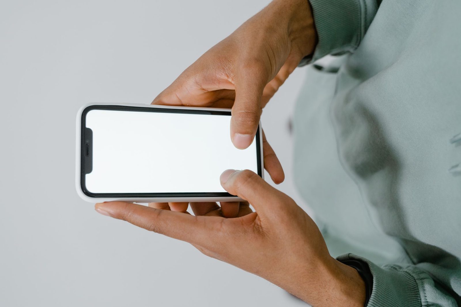 Mężczyzna trzymający smartfon poziomo w obu dłoniach, pusty biały ekran, na jasnoszarym tle