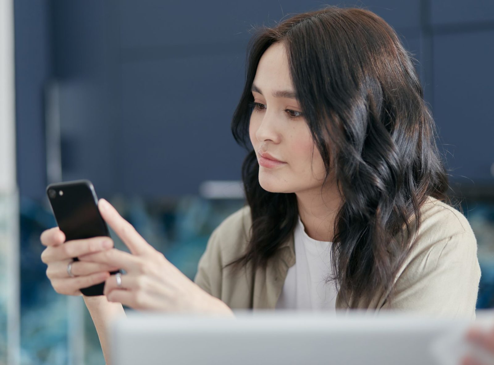 Giovane donna seduta davanti a un computer portatile, che guarda lo schermo dello smartphone tenuto con entrambe le mani
