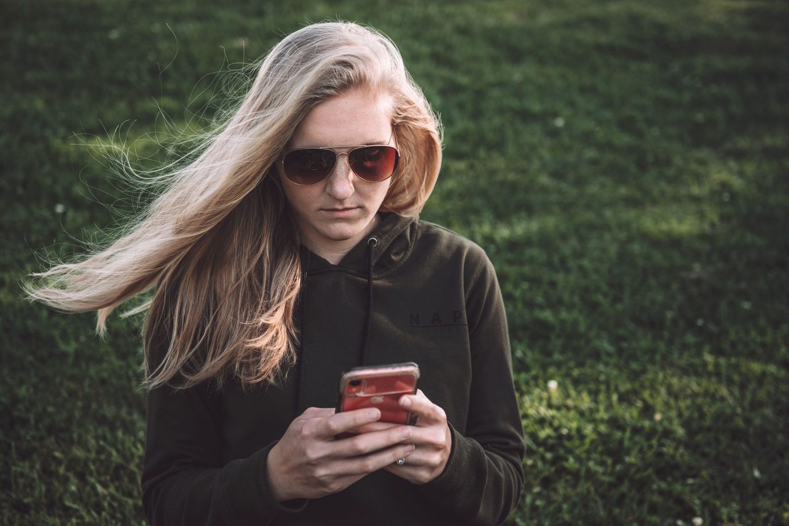 Giovane donna bionda con occhiali da sole che consulta lo smartphone all'aperto, su uno sfondo di erba verde