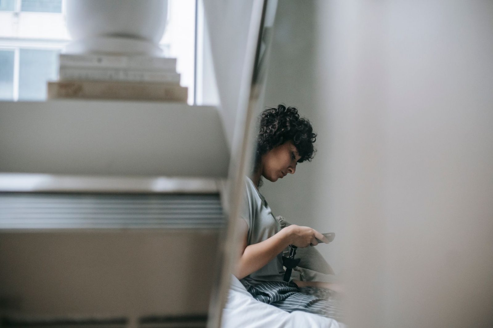 Donna seduta su un letto, con aria preoccupata, che consulta il telefono cellulare accanto a una scala