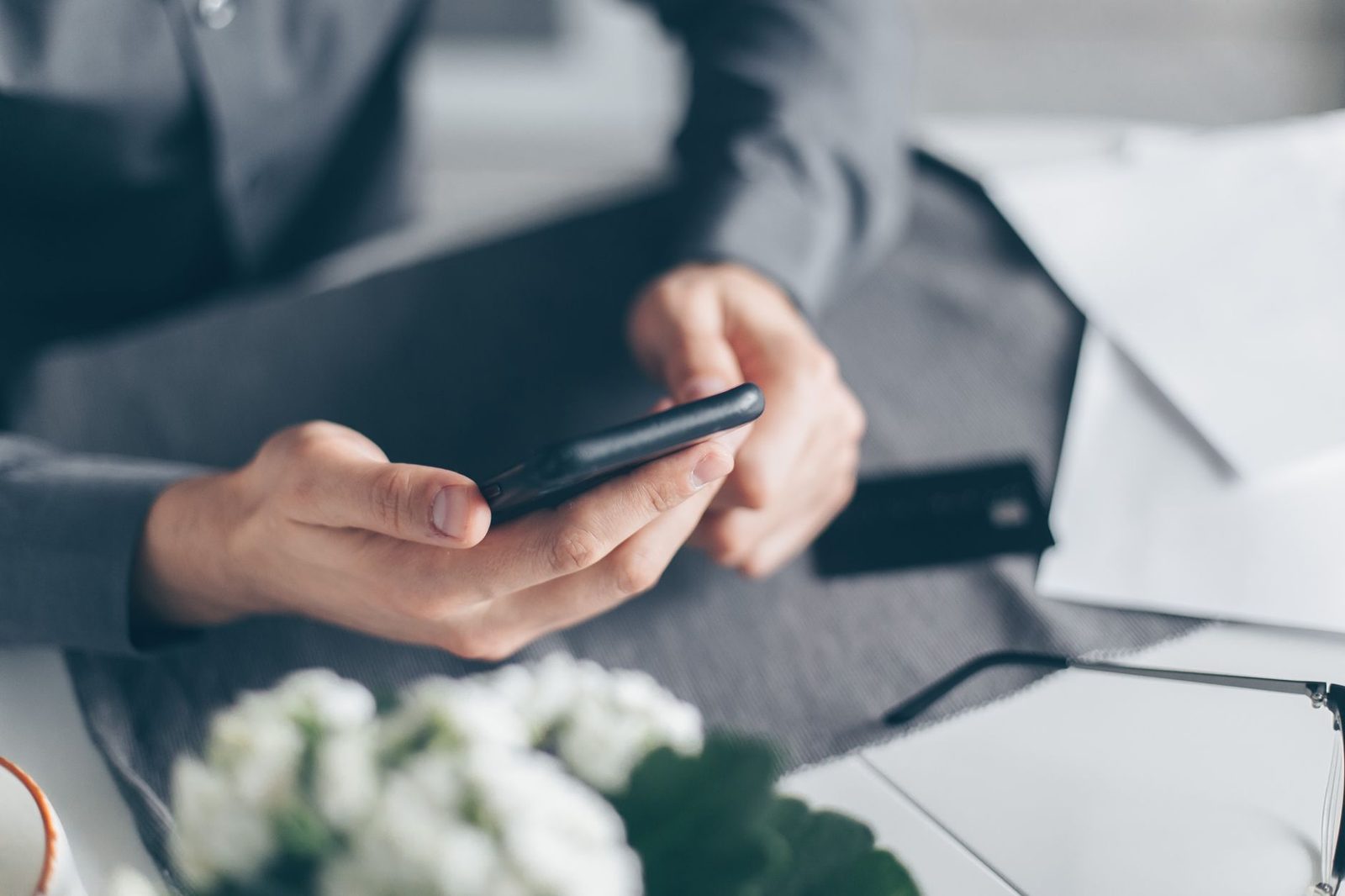 Mann im Anzug hält ein Smartphone in beiden Händen, daneben eine Bankkarte und Briefumschläge auf einem Schreibtisch