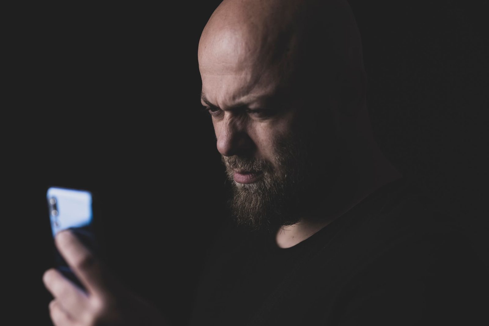 Bärtiger Mann mit rasiertem Kopf blickt im Dunkeln misstrauisch auf sein Smartphone-Display