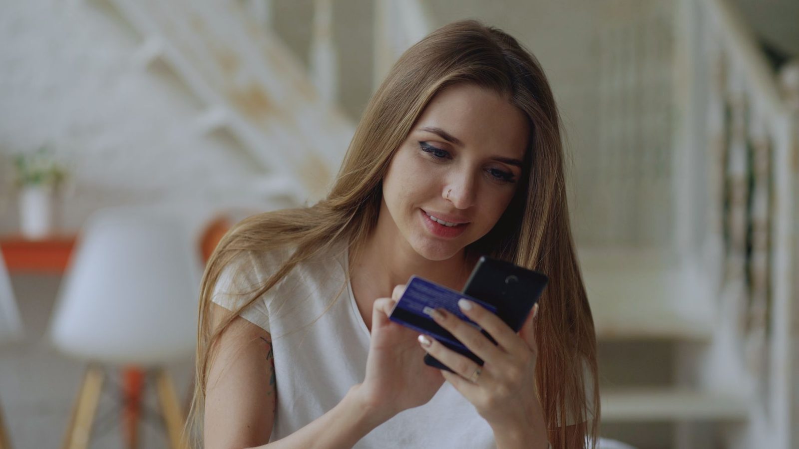 Jeune femme souriante tenant une carte bancaire et un smartphone dans son salon