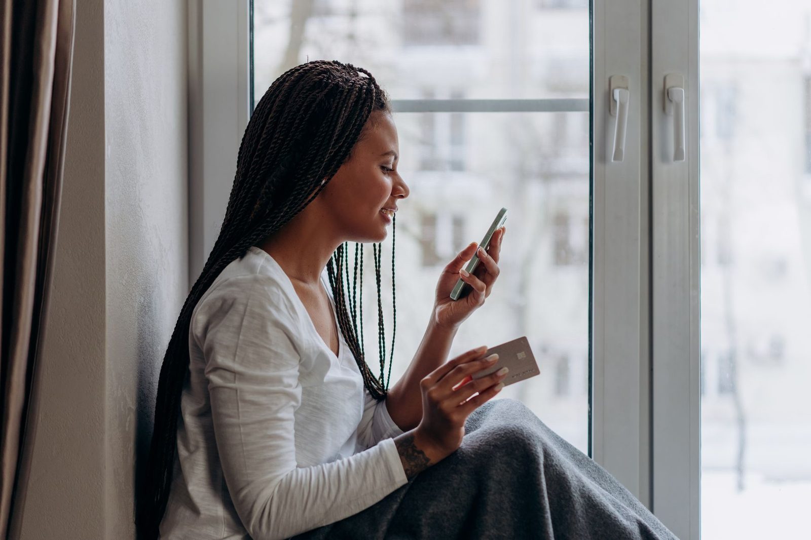 Femme assise près d'une fenêtre regardant son smartphone en tenant une carte bancaire à la main