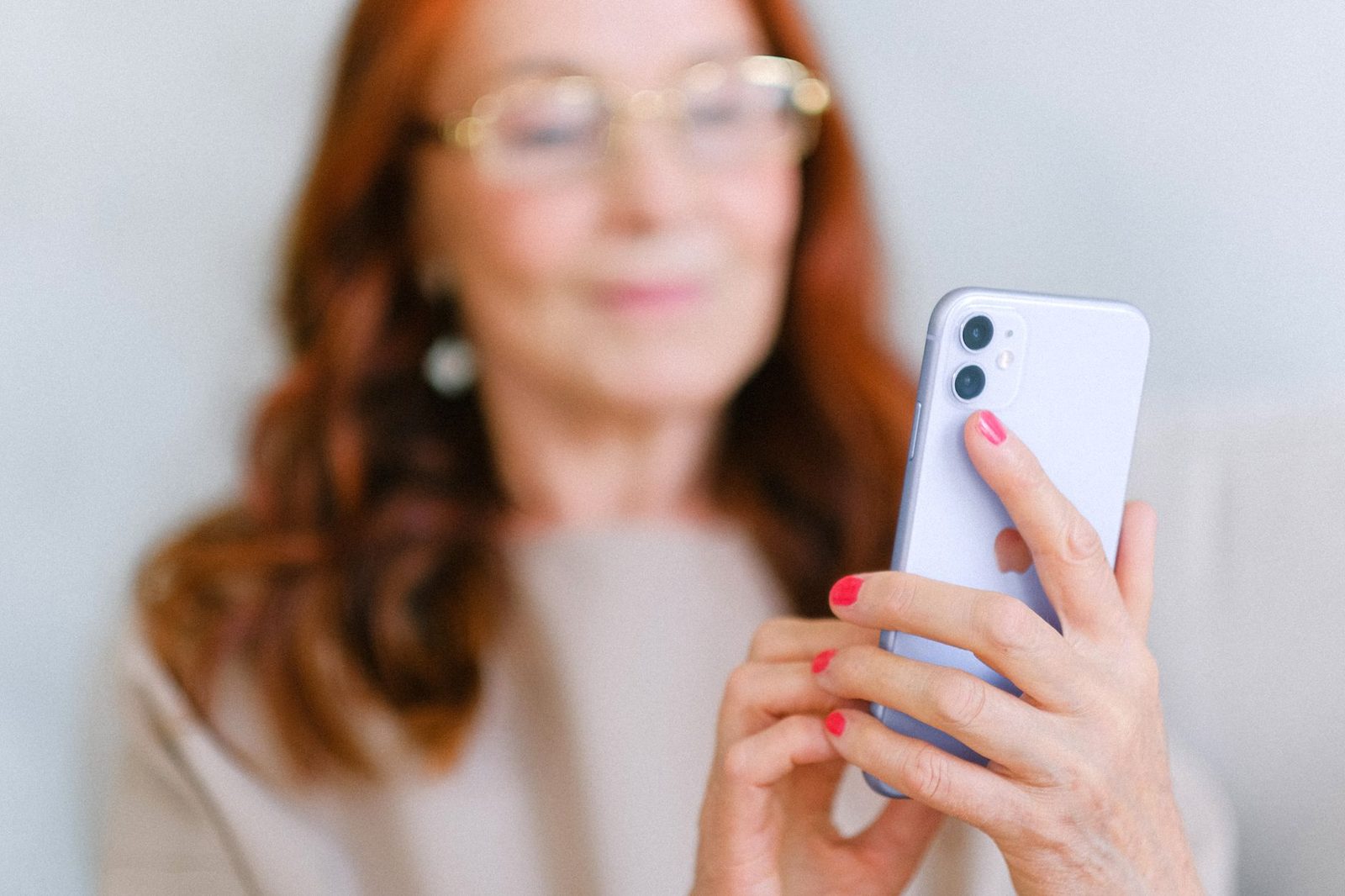 Femme aux cheveux roux et lunettes, floue en arrière-plan, tenant un smartphone blanc au premier plan