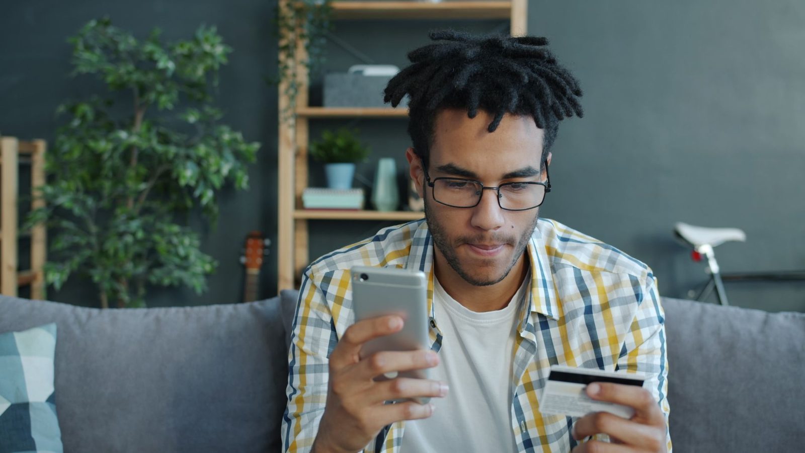Hombre con gafas sentado en un sofá mirando su smartphone con una tarjeta bancaria en la mano