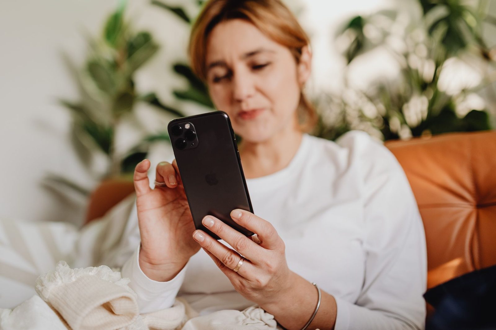 Frau auf einem Sofa, die mit beiden Händen ein Smartphone hält und darauf schaut, im Hintergrund Grünpflanzen