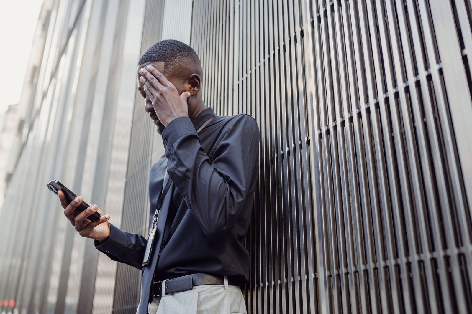 Hombre con camisa oscura, la mano en la cara, mirando su smartphone delante de una fachada metálica
