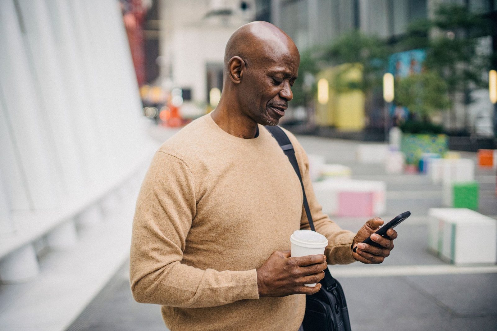Hombre con jersey beige consultando su teléfono móvil en la calle, con un vaso de café en la mano