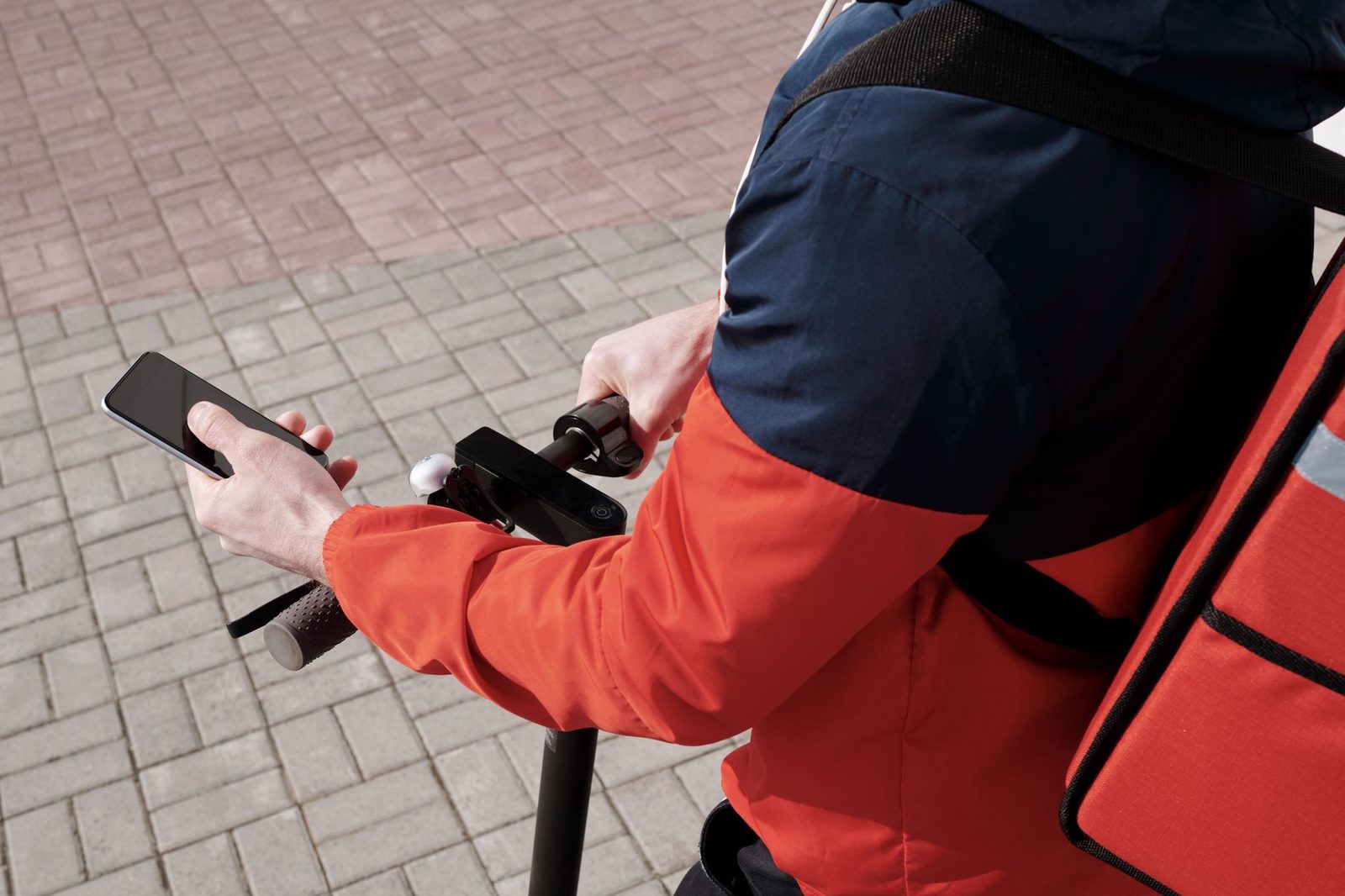 Fattorino con giubbotto arancione su un monopattino che guarda il suo smartphone con lo schermo spento