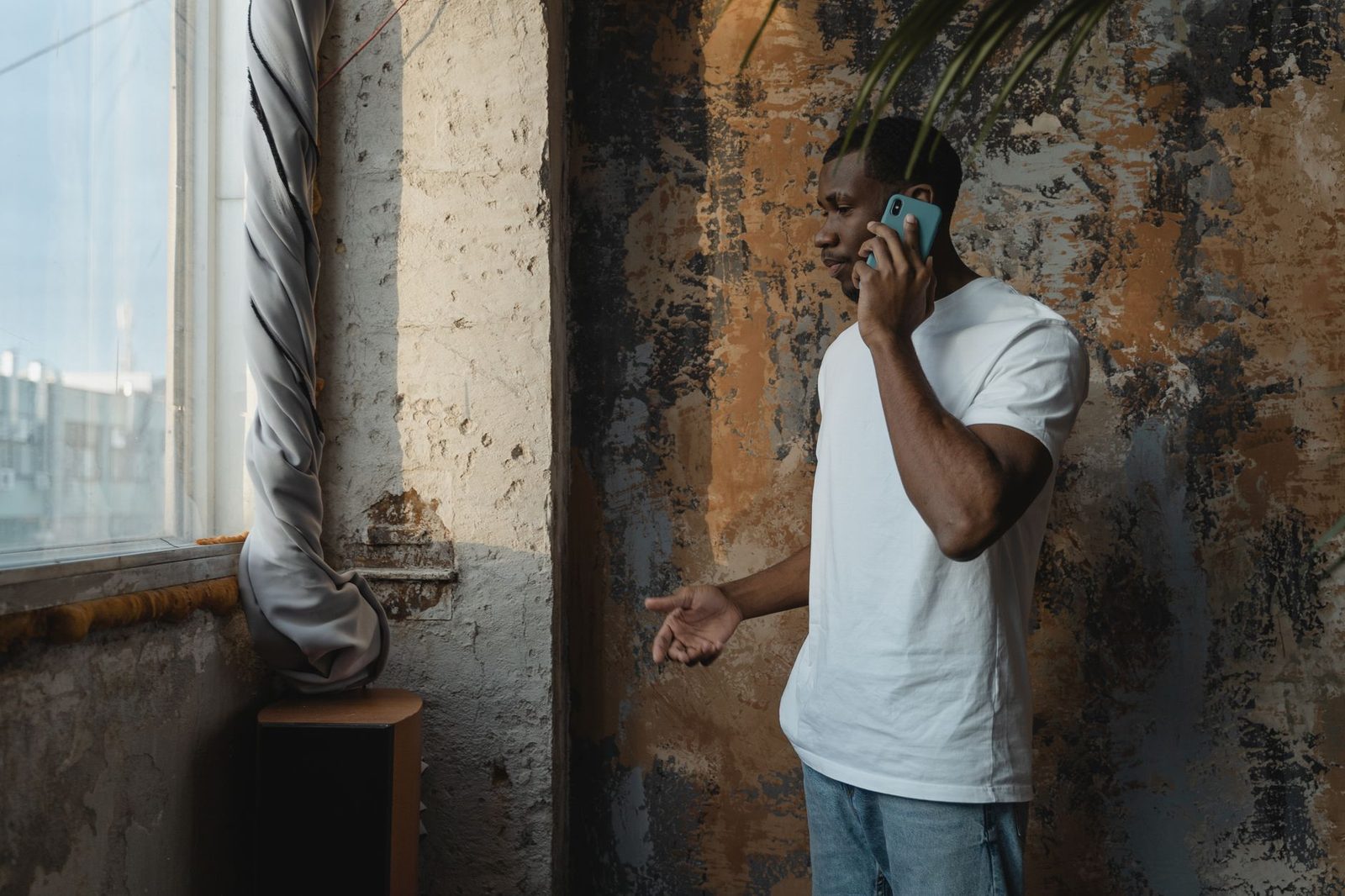 Uomo con t-shirt bianca al telefono vicino a una finestra, con aria perplessa