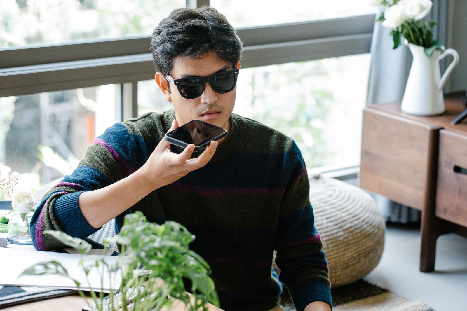Mann mit Sonnenbrille hält ein Smartphone nah ans Gesicht, sitzend am Fenster