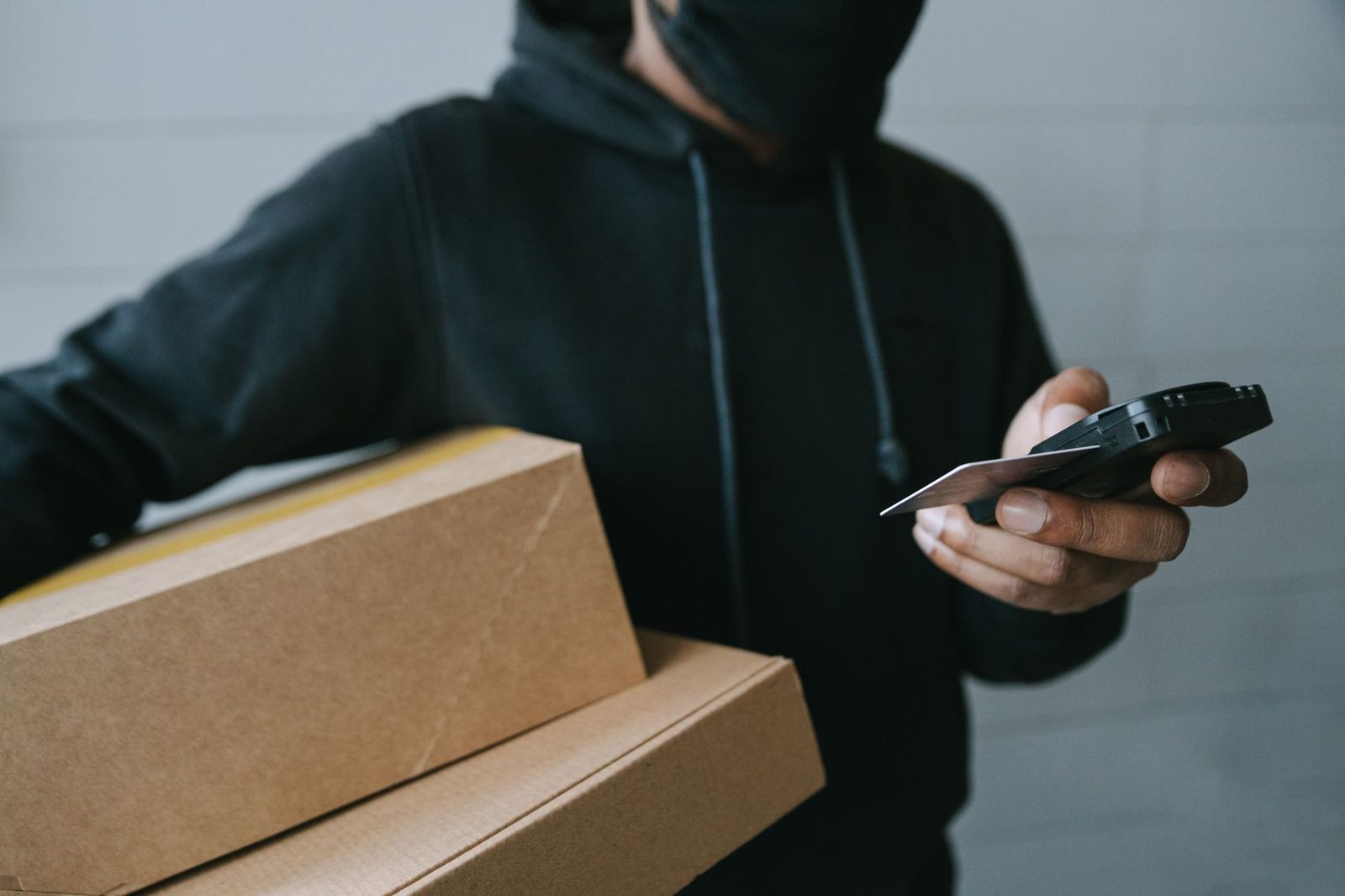 Mann mit Kapuze und Maske hält Pappkartons, ein Smartphone und eine Bankkarte in der Hand