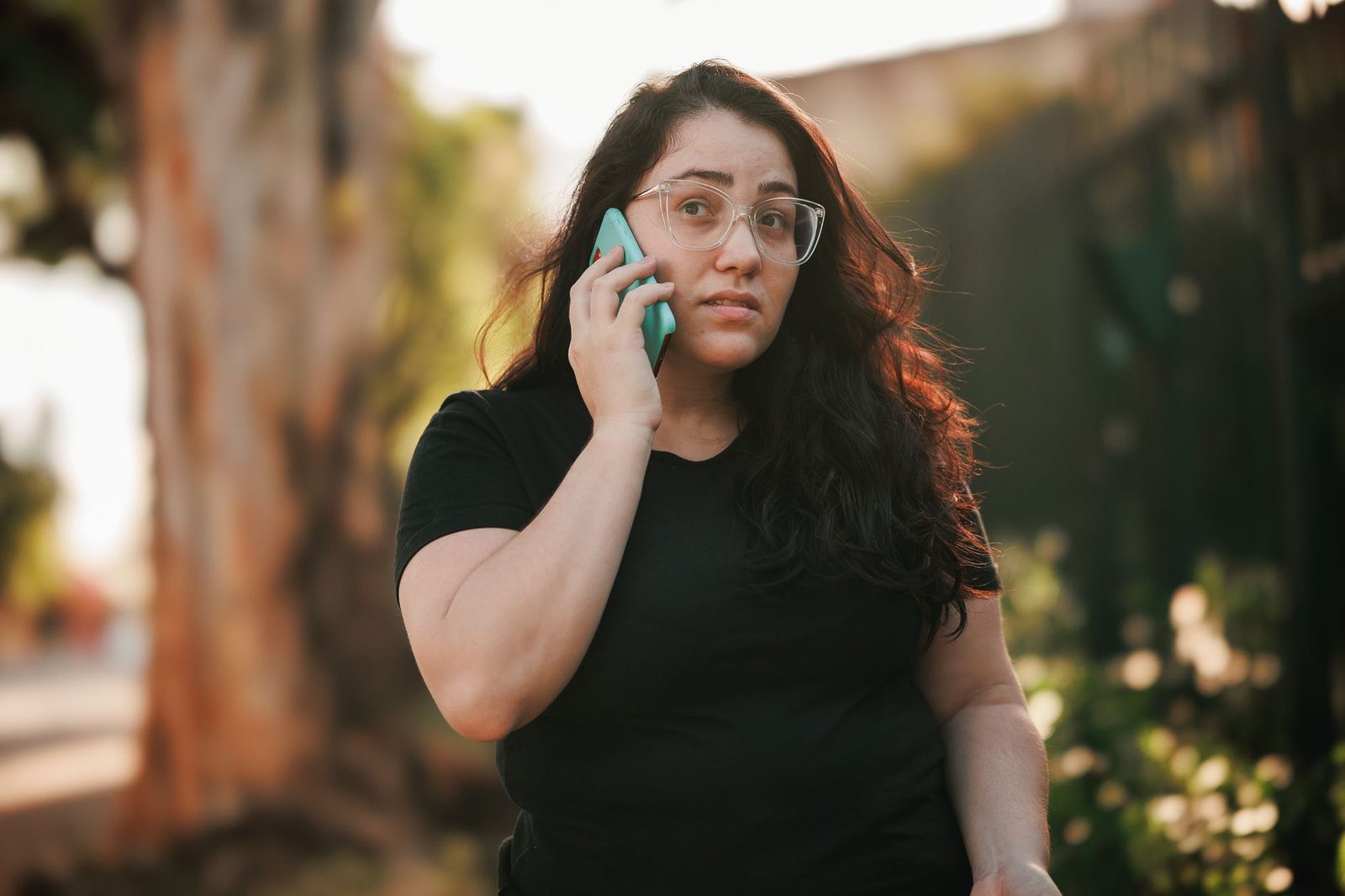 Young woman wearing glasses, looking worried, talking on a turquoise smartphone outdoors