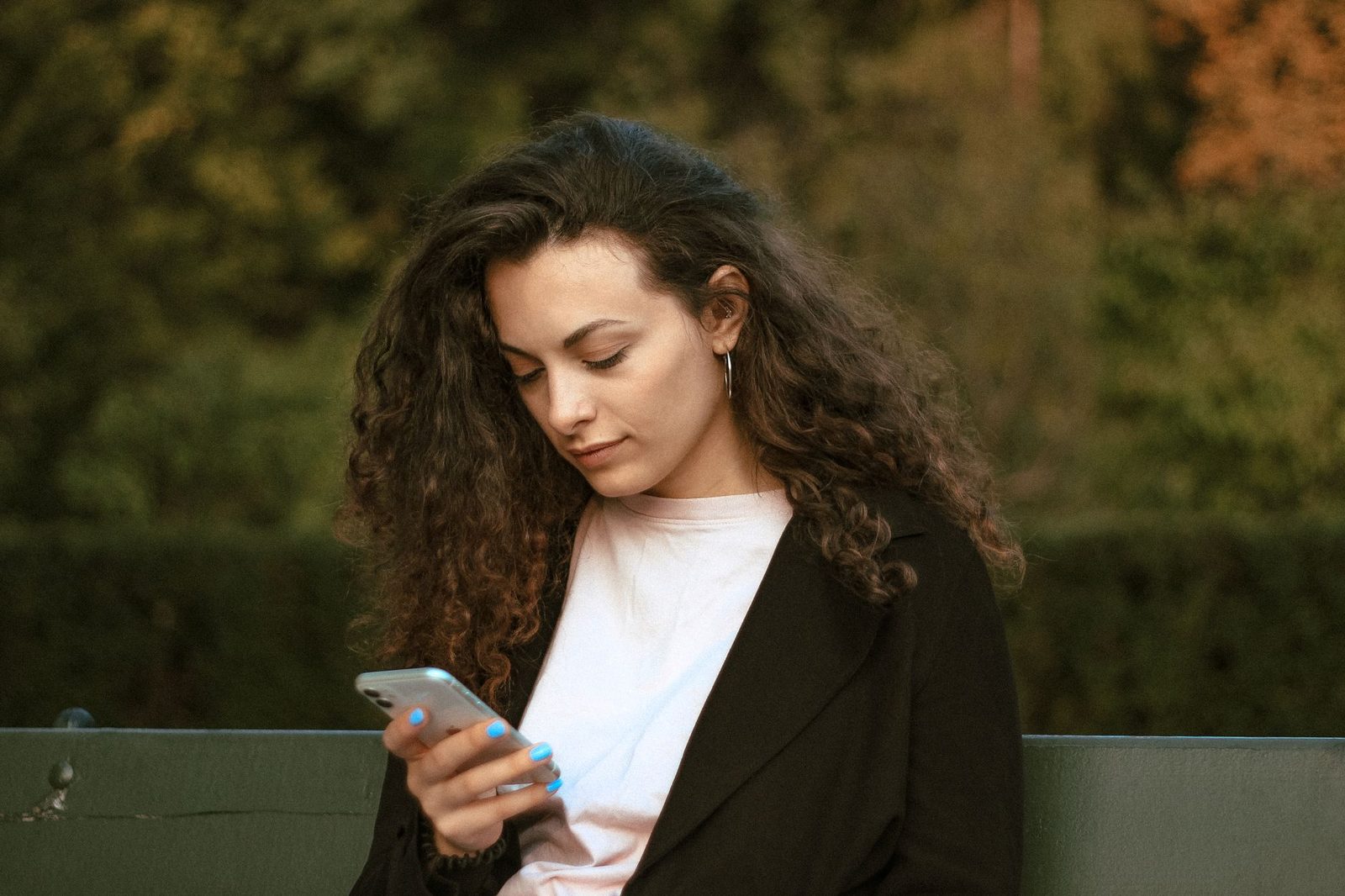 Mujer joven de pelo rizado sentada en un banco, consultando su smartphone en un parque