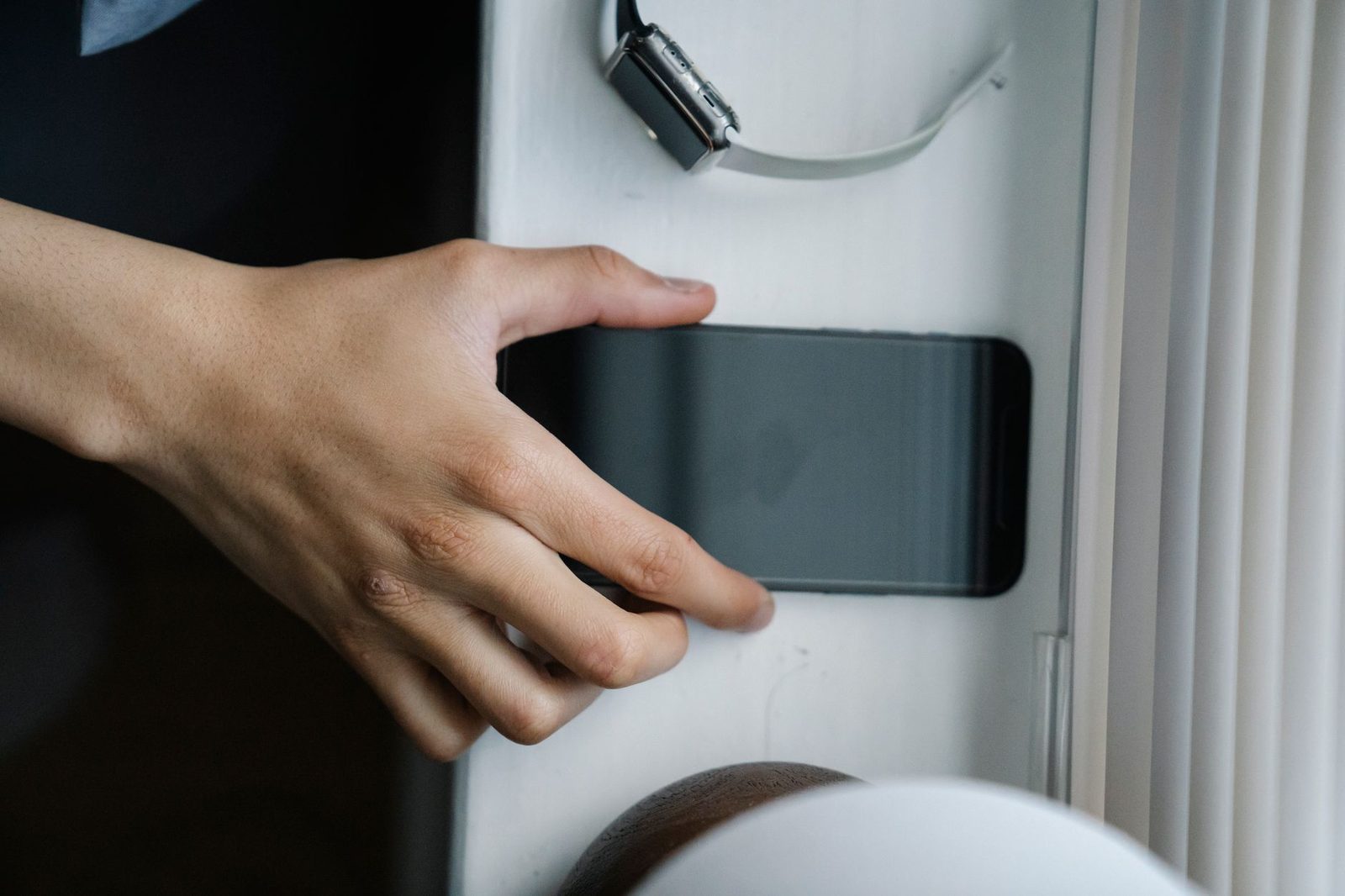 Une main saisit un smartphone à l'écran éteint posé sur une table blanche, près d'une montre connectée