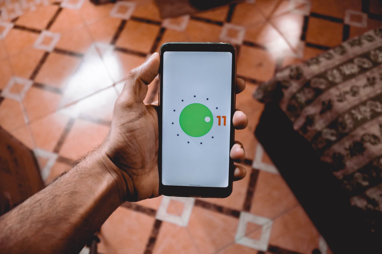 Main tenant un smartphone Android affichant le logo de démarrage Android 11, au-dessus d'un sol carrelé