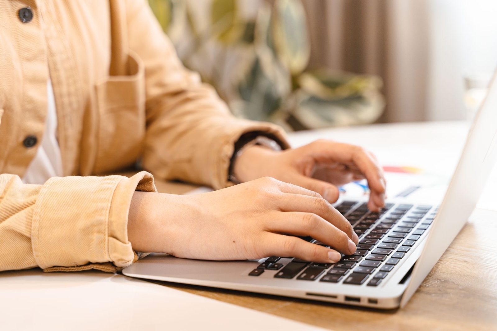Close-up of a woman's hands in a beige jacket typing on a laptop keyboard