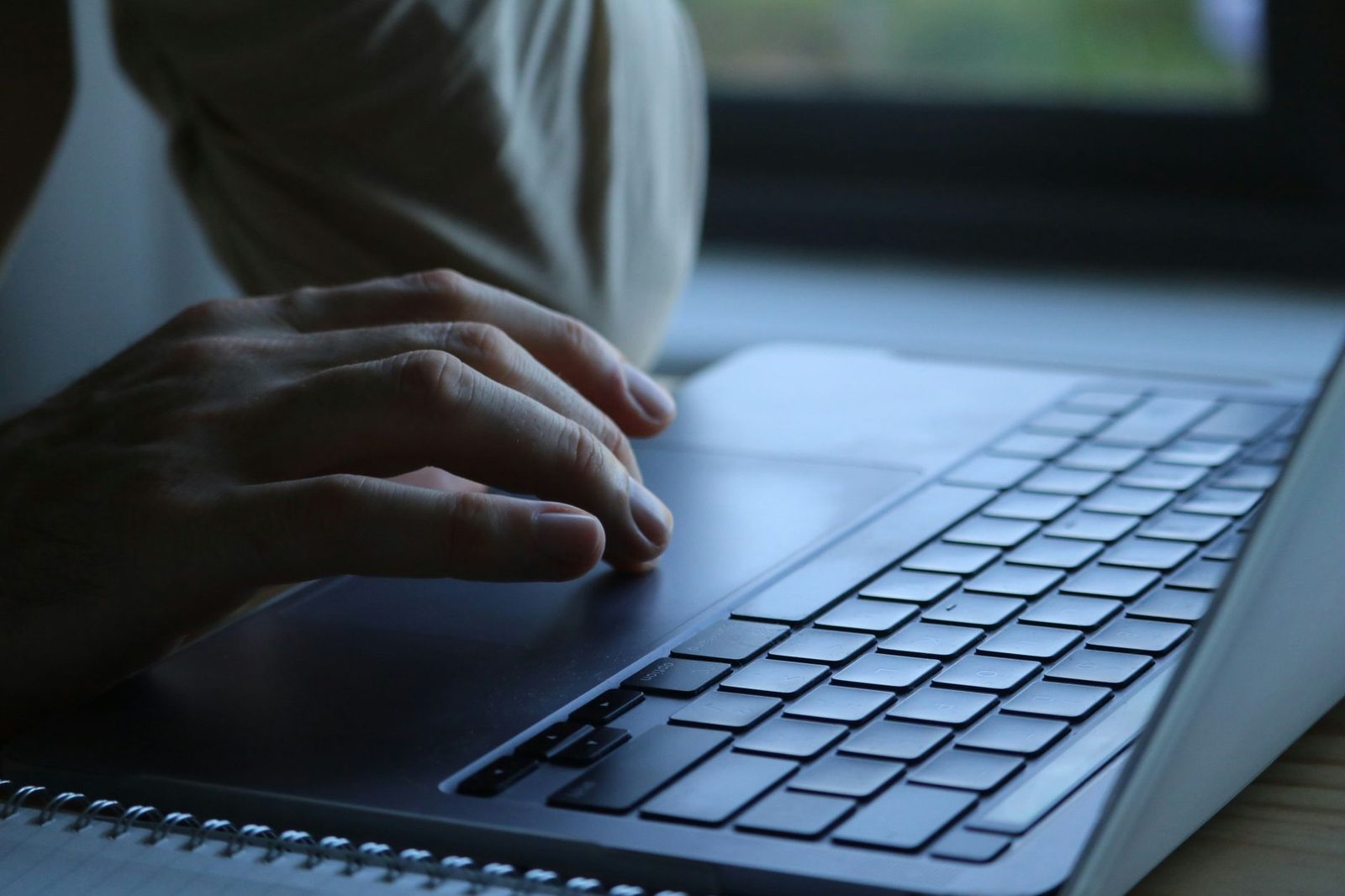 Hand resting on a laptop touchpad, keyboard in the foreground near a window