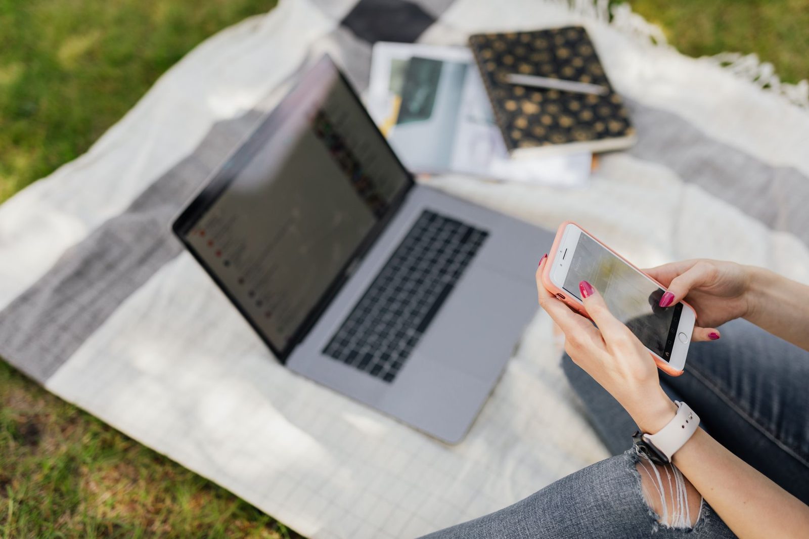 Femme assise sur un plaid dans l'herbe tenant un smartphone devant un ordinateur portable ouvert