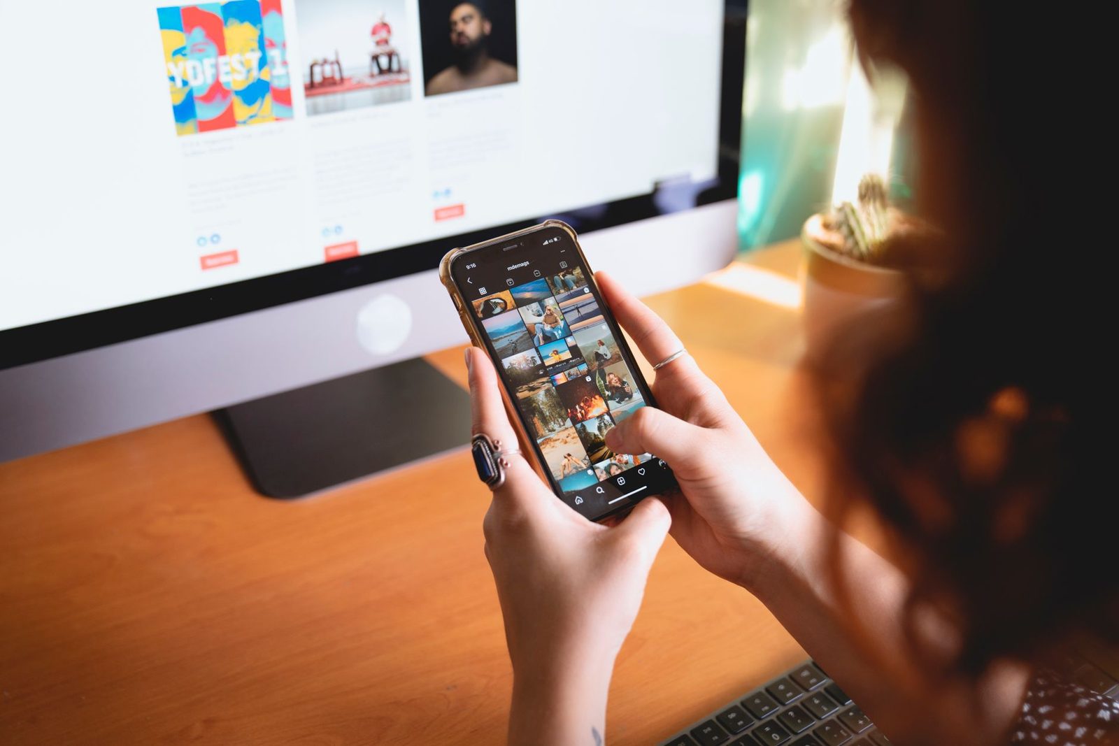 Femme tenant un smartphone devant un ordinateur de bureau allumé, sur un bureau en bois