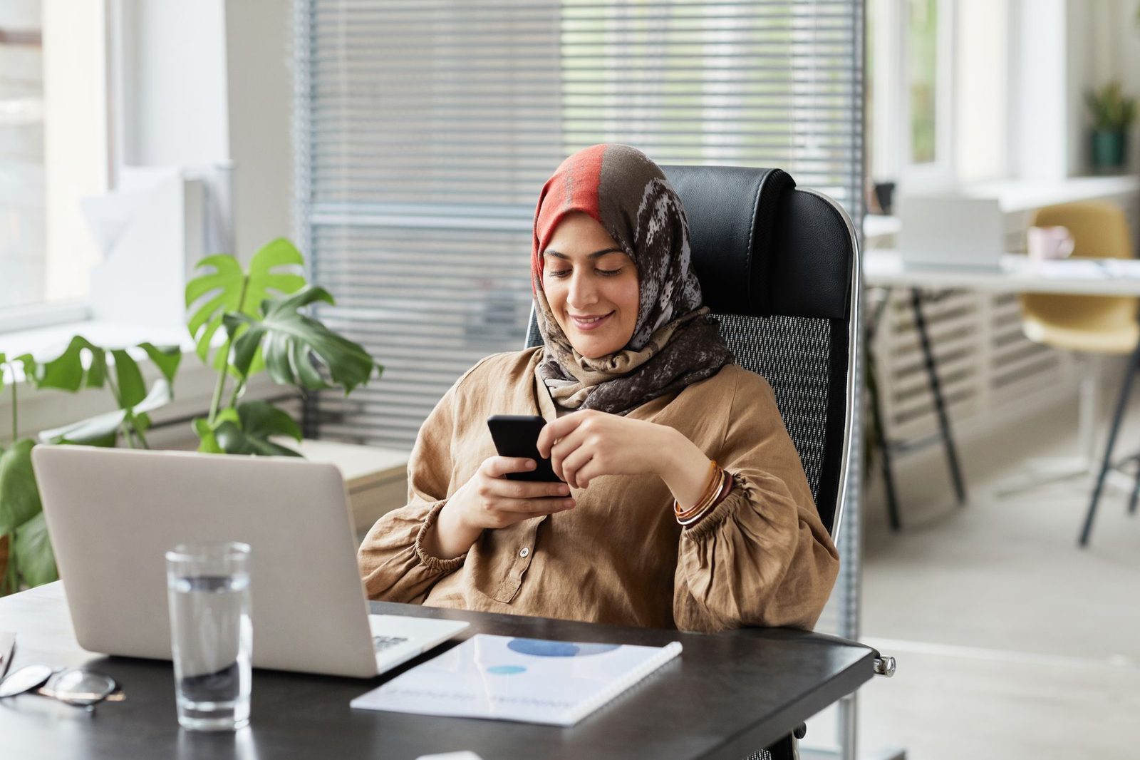 Smiling woman at her desk sending a message on her smartphone in front of an open laptop